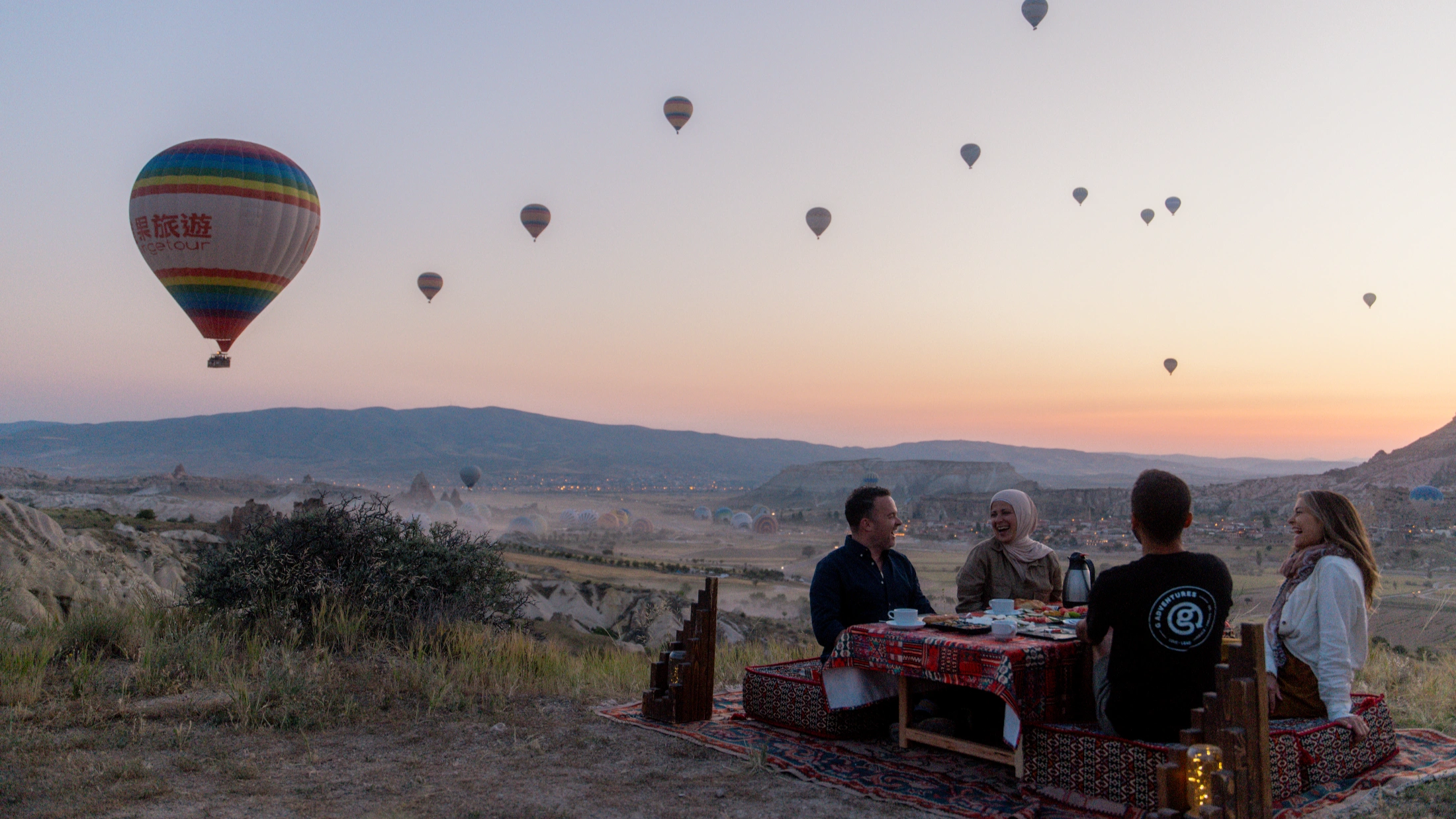 Four people enjoy an outdoor breakfast in Cappadocia, Turkey, with colorful hot air balloons filling the pastel sunrise sky. Four people enjoy an outdoor breakfast in Cappadocia, Turkey, surrounded by colorful hot air balloons at sunrise.