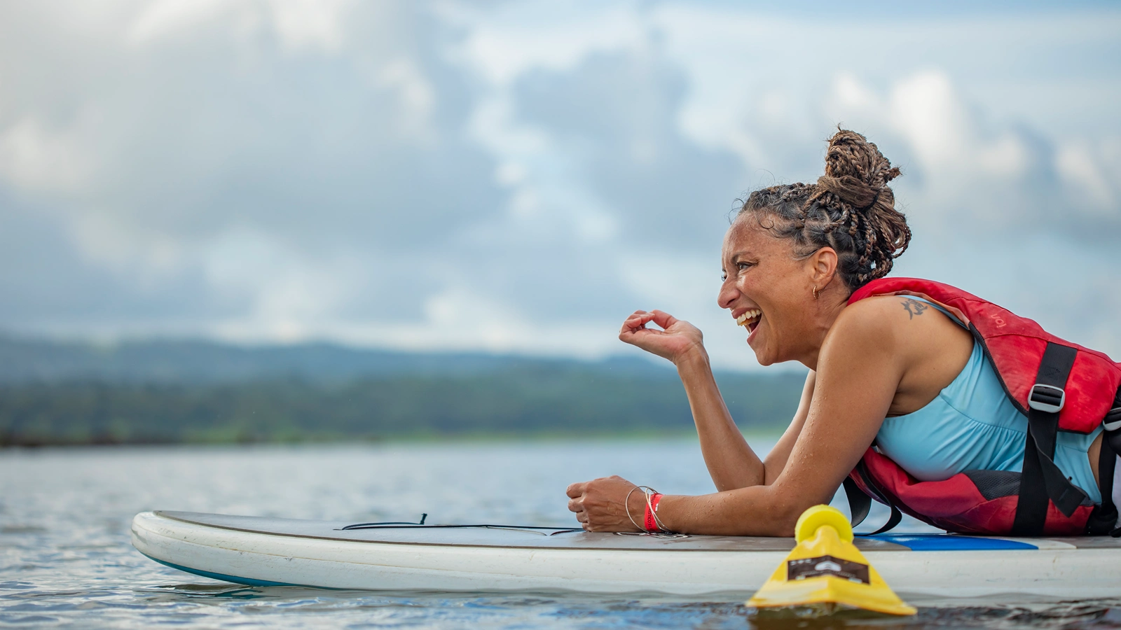 A laughing woman in a blue top and red life vest lies on a paddleboard on calm water with cloudy skies behind her. A laughing woman in a blue top and red life vest lies on a paddleboard on calm water with cloudy skies behind her.