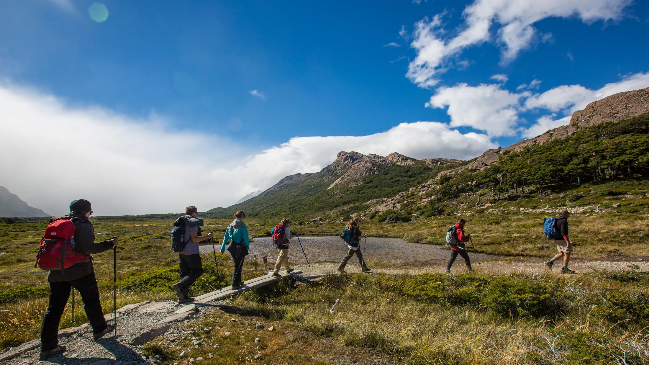 Group of hikers with backpacks and trekking poles crossing a wooden bridge in a green mountain valley under a blue sky. Group of hikers with backpacks and trekking poles crossing a wooden bridge in a green mountain valley under a blue sky.