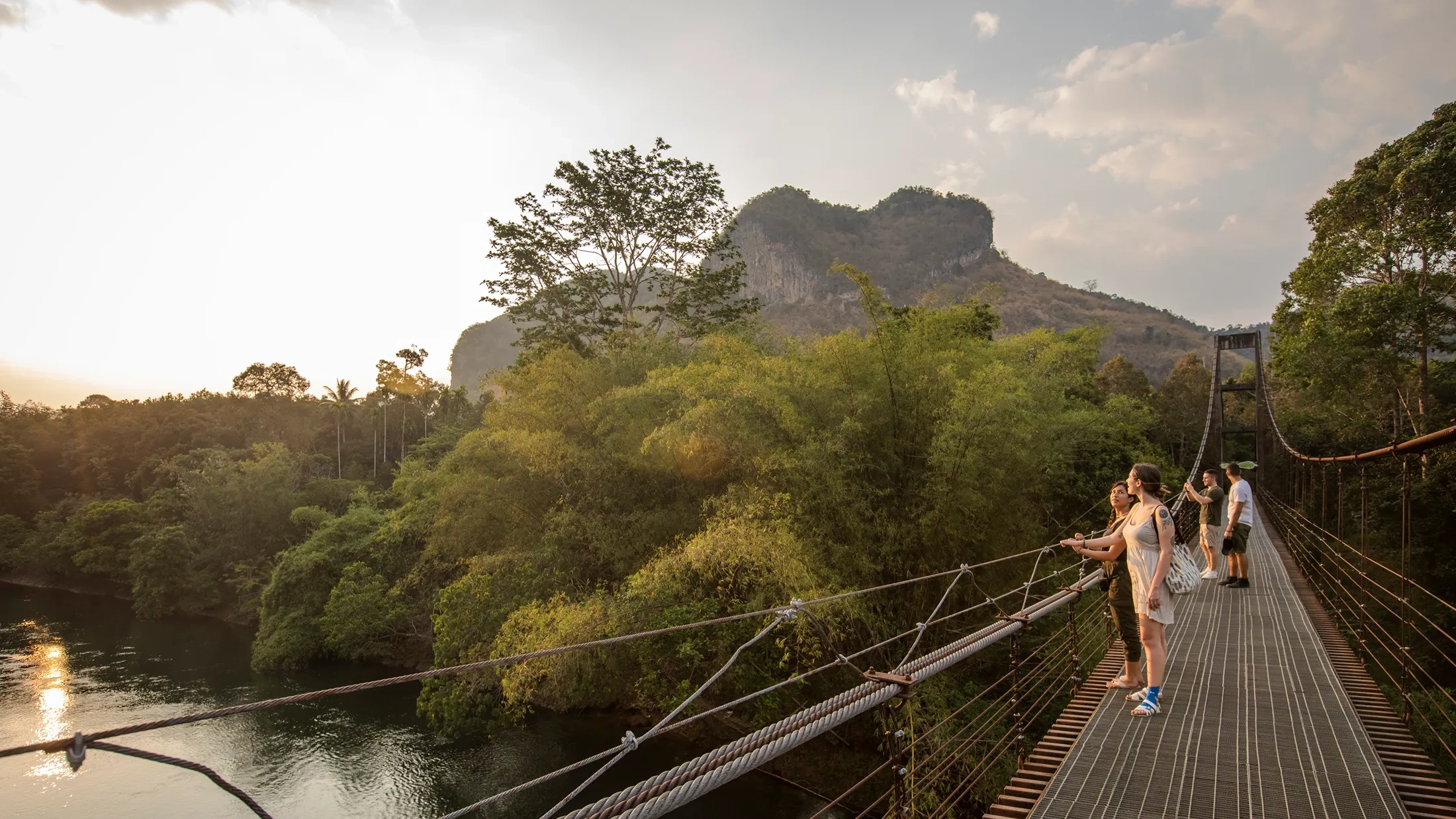 Tourists stand on a suspension bridge over a river, surrounded by lush green trees and a rocky mountain at sunset. Tourists stand on a suspension bridge over a river, surrounded by lush green trees and a rocky mountain at sunset.