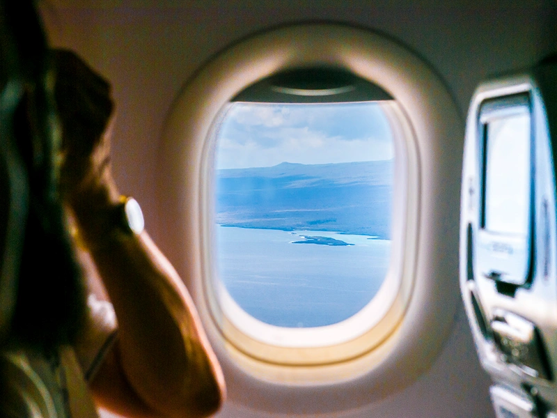 View from airplane cabin window showing blue ocean, small island, and mountains under a partly cloudy sky. View from airplane cabin window showing blue ocean, small island, and mountains under a partly cloudy sky.