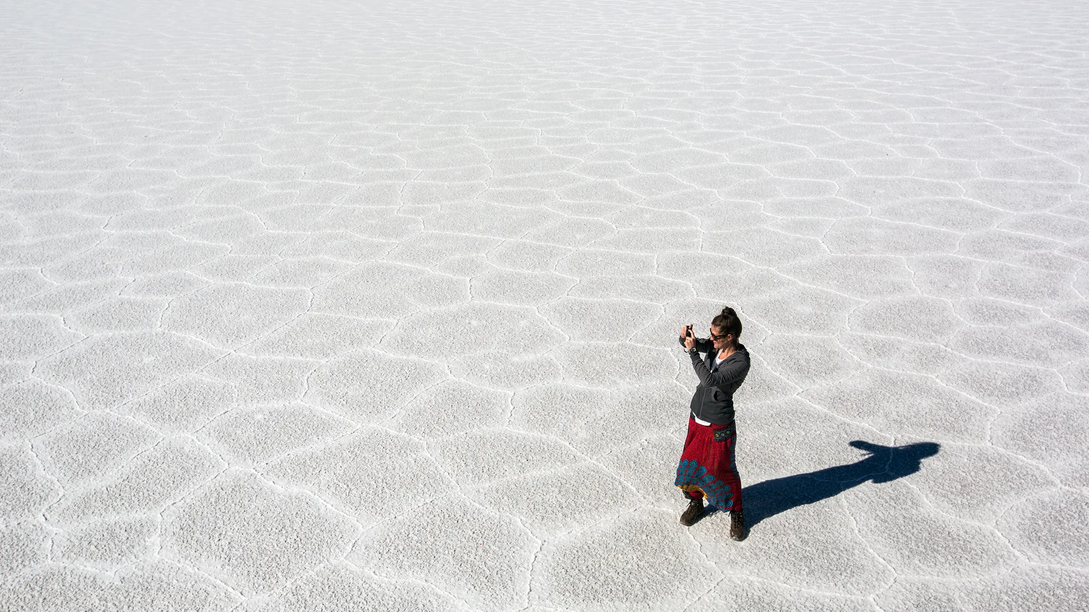 A woman in a red skirt photographs the vast white salt flats, with hexagonal salt patterns stretching across the landscape. A woman in a red skirt photographs the vast white salt flats, with hexagonal salt patterns stretching across the ground.
