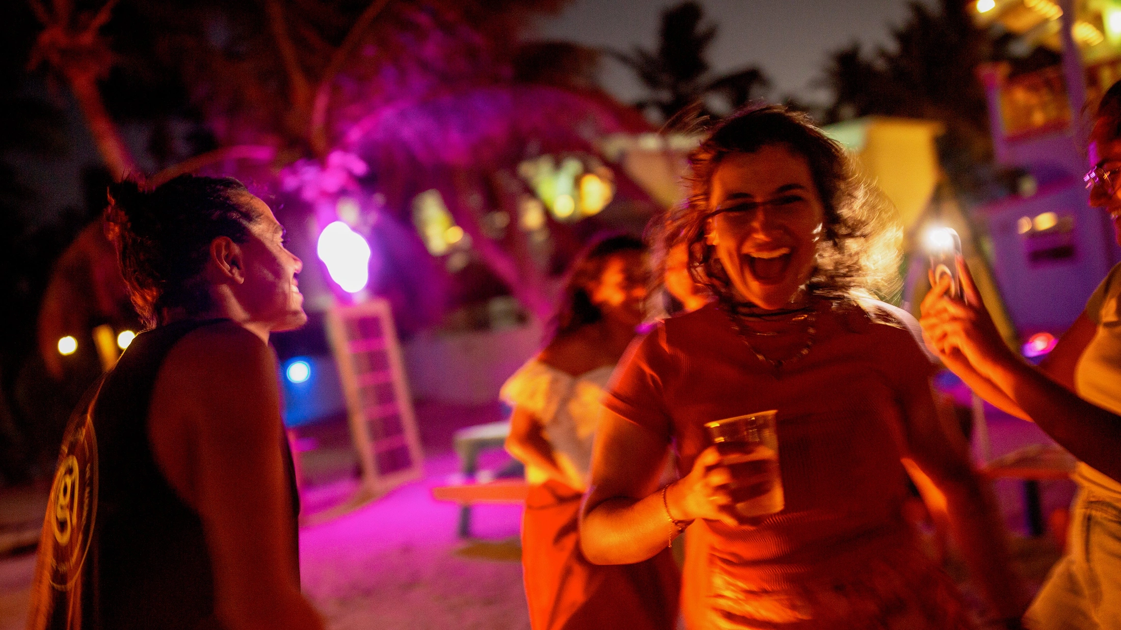 People laughing and dancing at an outdoor night party with colorful pink and orange lights, one holding a drink. People laughing and dancing at an outdoor night party with colorful pink and orange lights, one holding a drink.