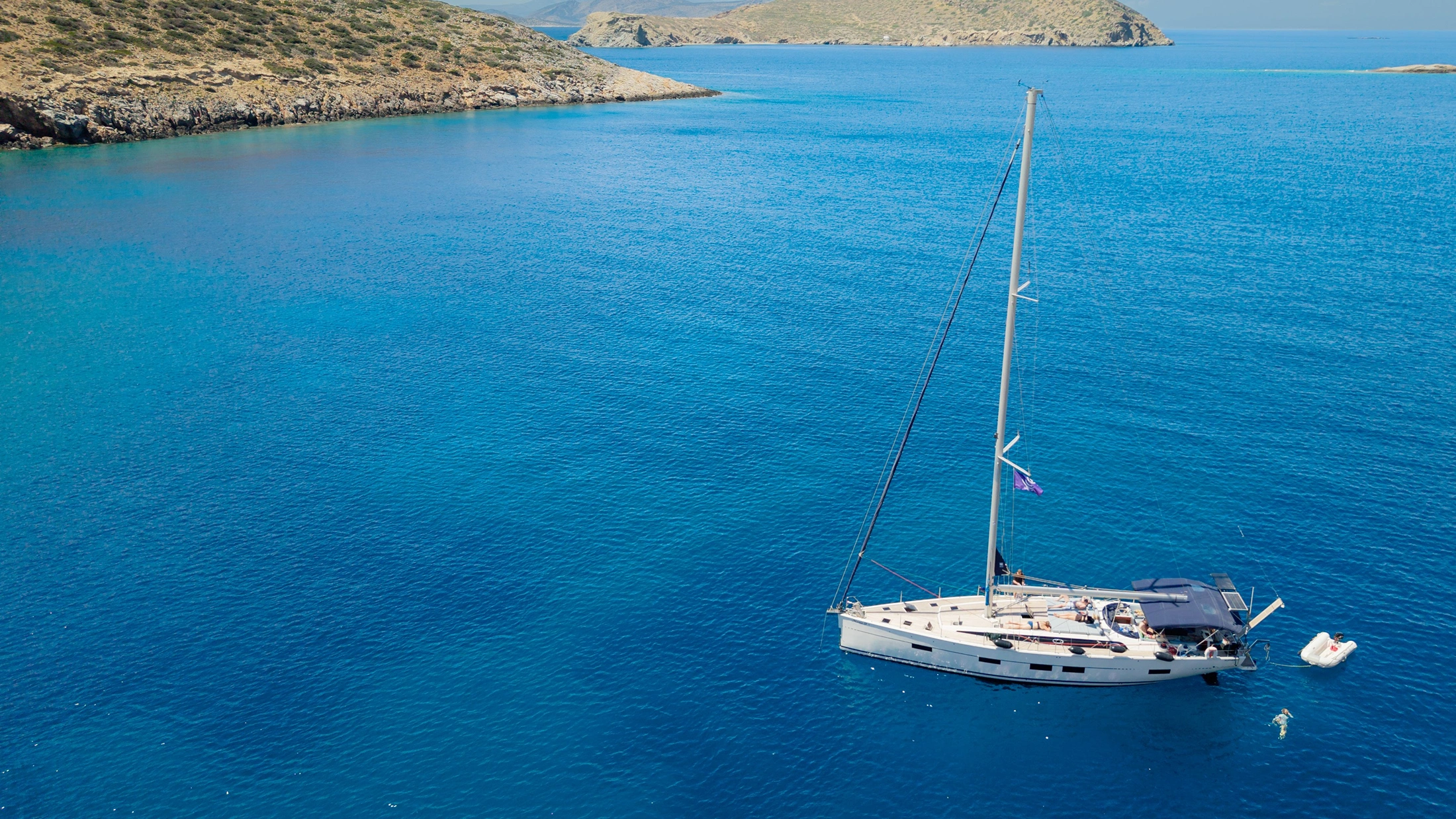 Aerial view of a white sailboat anchored in calm blue waters near rocky islands, with a small dinghy and swimmer nearby. Aerial view of a white sailboat anchored in calm blue waters near rocky islands, with a small dinghy and swimmer nearby.