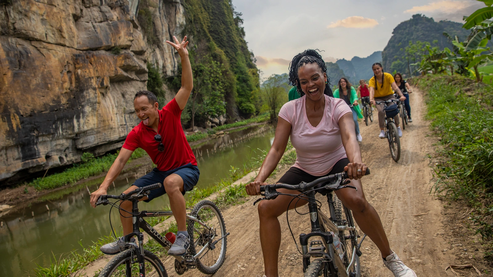 Clone A laughing woman in a blue top and red life vest lies on a paddleboard on calm water with cloudy skies behind her. A joyful group cycling on a dirt path beside a river, surrounded by limestone cliffs and lush greenery.