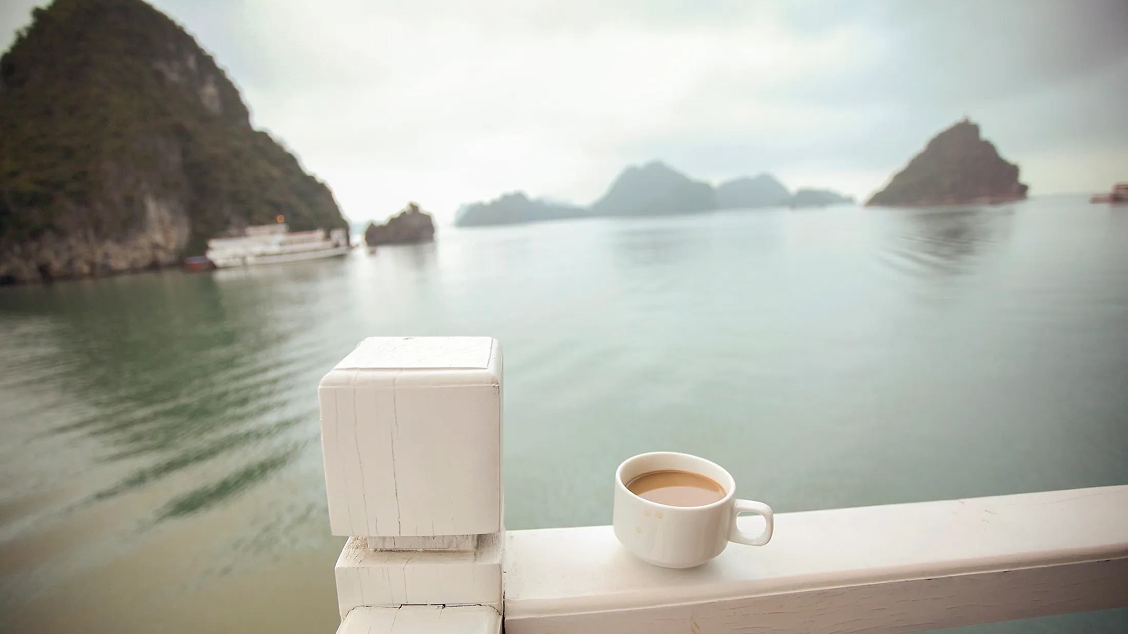A white coffee cup on a railing overlooks a calm bay with misty limestone islands and a boat under overcast skies. A white coffee cup on a railing overlooks a calm bay with misty limestone islands and a boat in the background.
