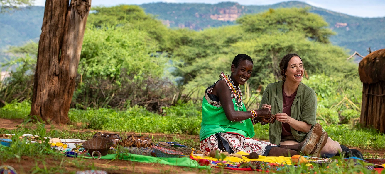 Two women laugh while beading outdoors in an African village, surrounded by colorful crafts, lush green trees, and hills. Two women laugh while beading outdoors in an African village, surrounded by colorful crafts, lush green trees, and hills.