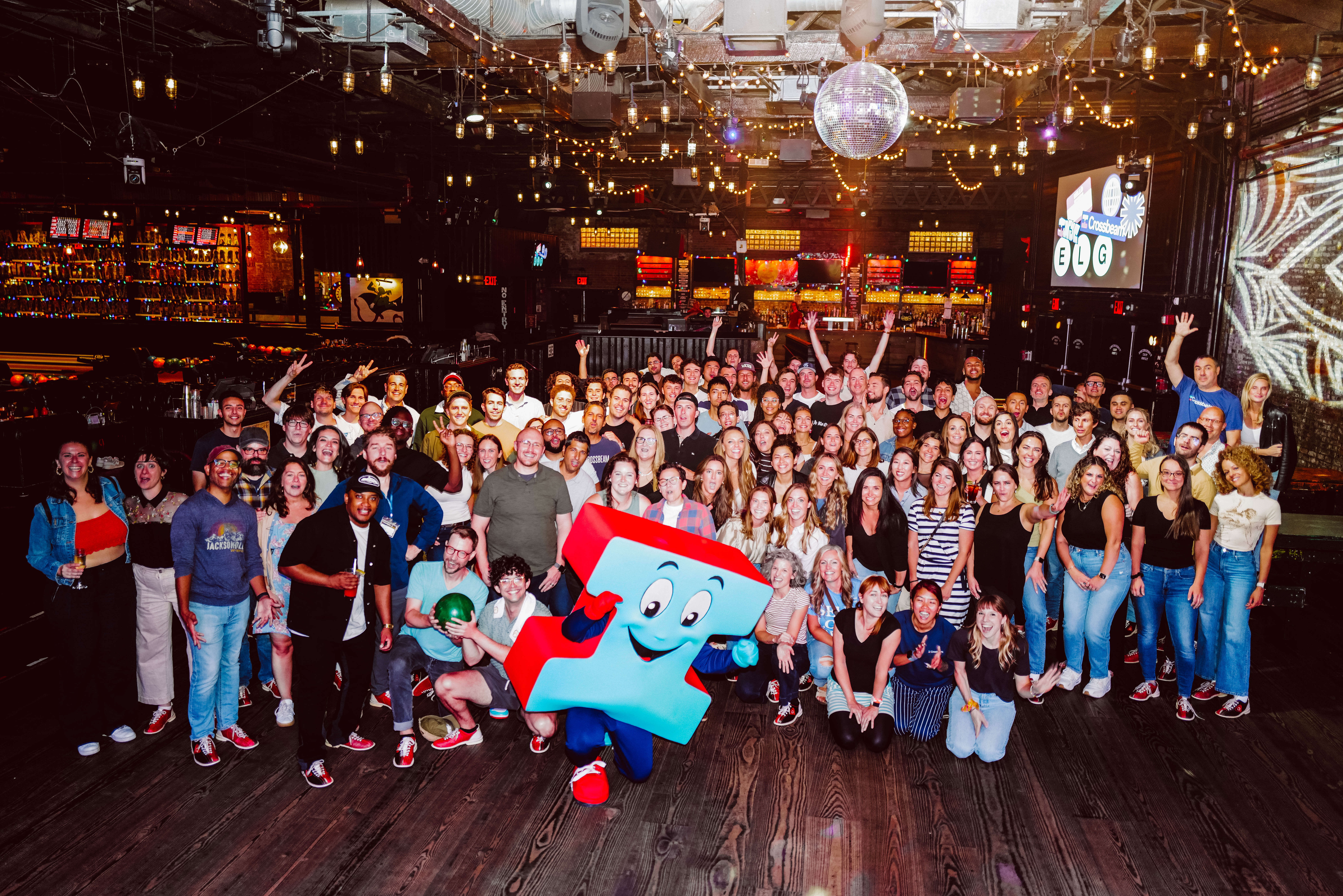 Crossbeam A large group of people pose together indoors, under festive lighting and decor that celebrates employee well-being benefits. Some hold drinks, with a person in a colorful mascot costume front and center. A bar and stage with equipment are visible in the background, adding to the joyful atmosphere.