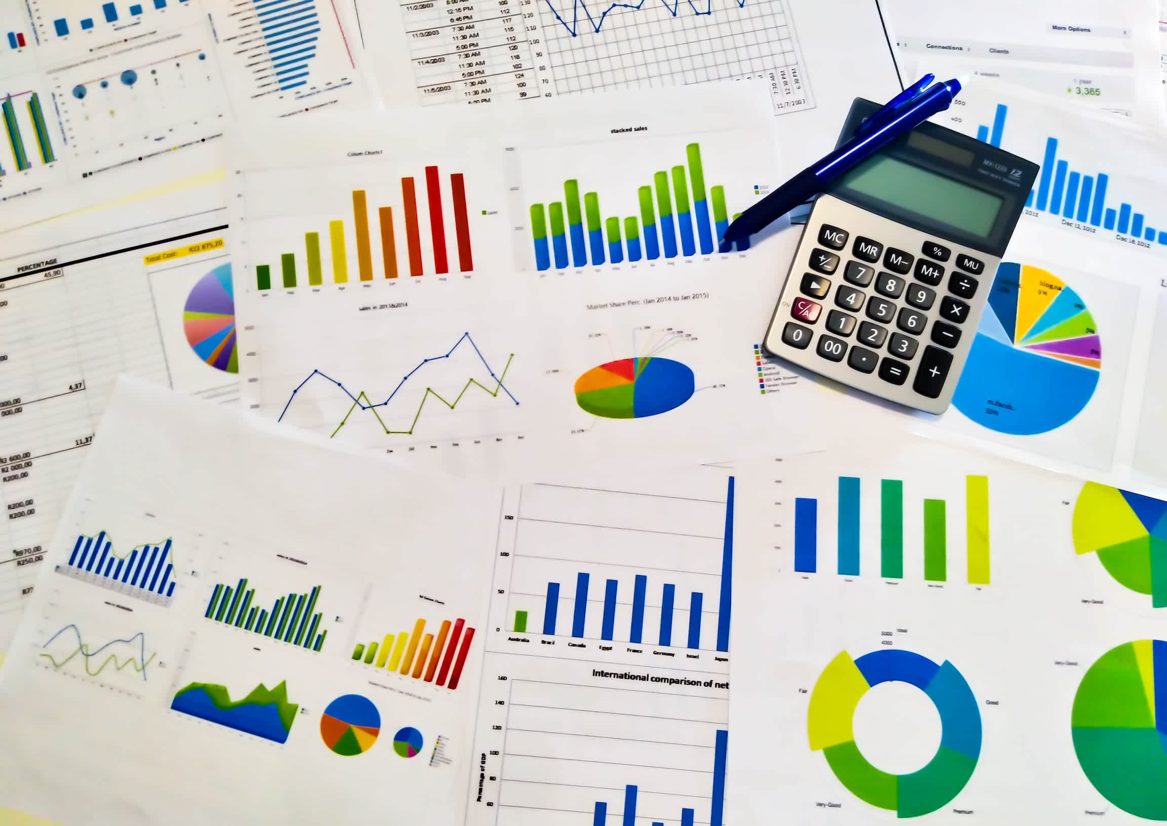 Photo full of spreadsheets and statistics charts spread across a surface with a calculator and pen nearby.