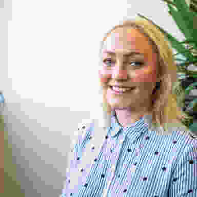 Art Director Headshot, mittelblondes Haar mit blauem Hemd, weißer Hintergrund, neben einer Pflanze.