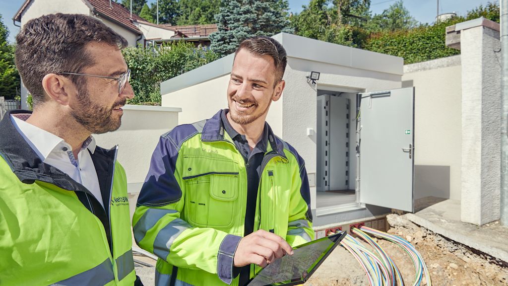Zwei Techniker vor einem Technikgebäude, besprechen Projekt am Tablet.