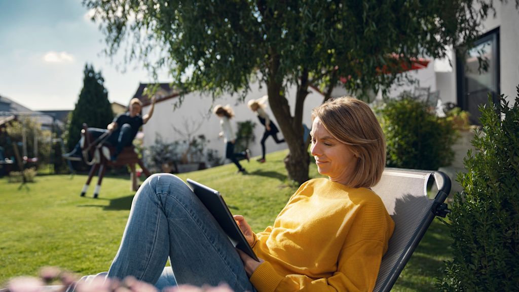 Frau entspannt im Garten, Tablet in der Hand. Kinder spielen im Hintergrund.