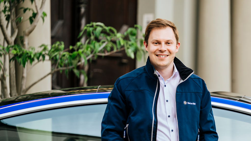 Mann in dunkler Jacke lächelt vor Auto.