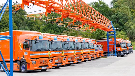 Reihe von orangefarbenen E-Lkw der Spedition Rüdinger unter einer auffälligen Overhead-Ladeinfrastruktur mit sieben Ladepunkten