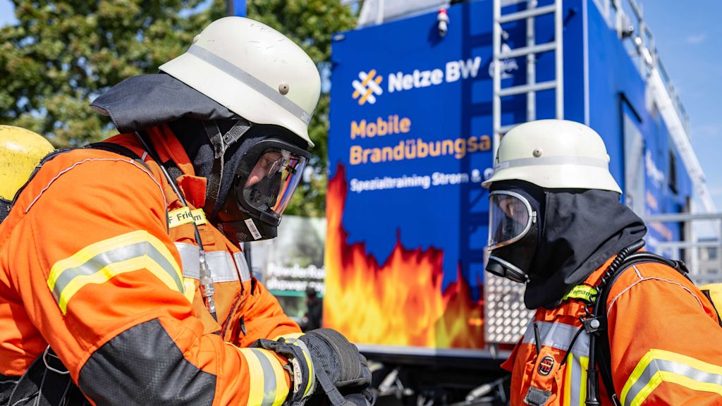 Zwei Feuerwehrleute in Schutzkleidung vor einem Brandübungswagen.