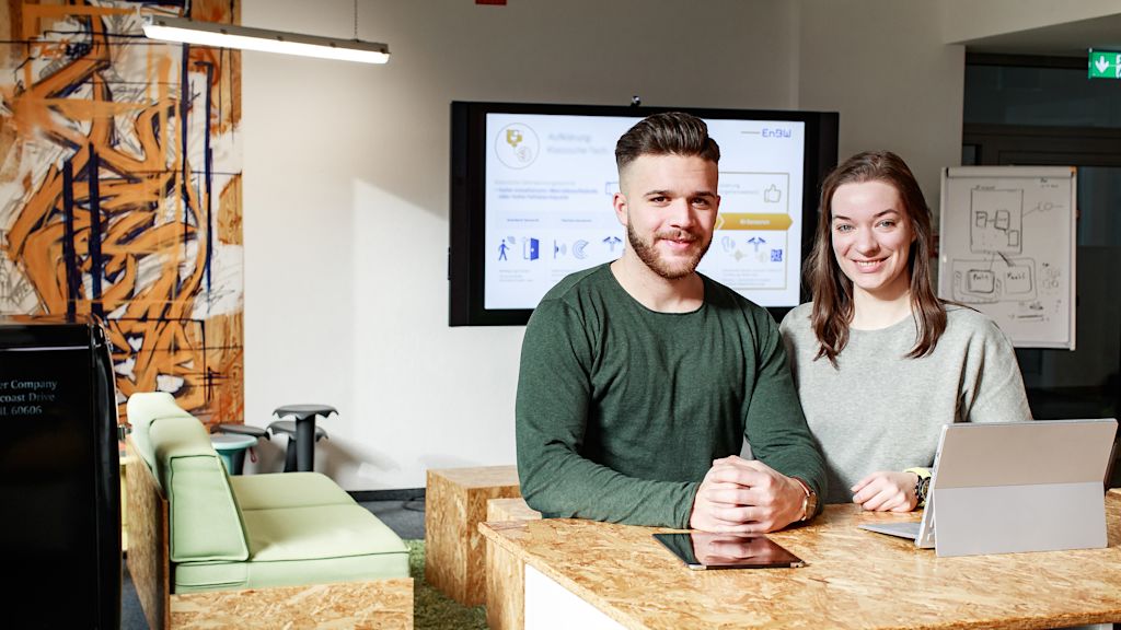 Zwei junge Leute sitzen vor einem Tablet und lächeln in die Kamera.