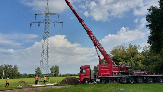Ein roter Kran hebt Arbeiter*innen zu einem hohen Strommast, während mehrere Personen in Warnwesten auf dem Boden stehen und arbeiten.