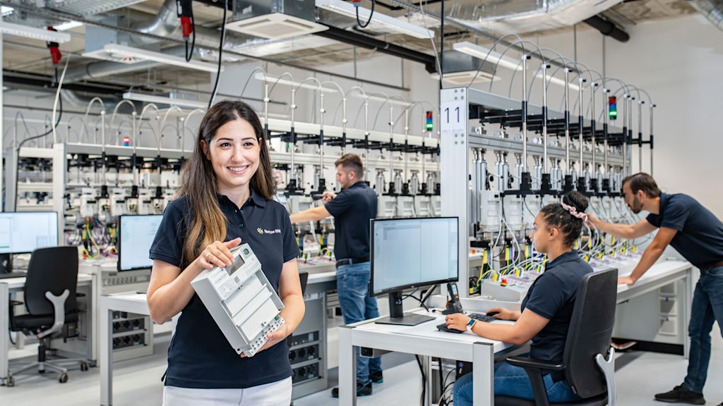 Mitarbeiterin der Netze BW GmbH hält einen digitalen Zähler in der Hand. Im Hintergrund ist ein Technikraum des Ausbildungszentrums zu sehen.