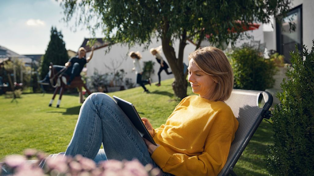 Frau entspannt im Garten, guckt aufs Tablet. Kinder spielen im Hintergrund.
