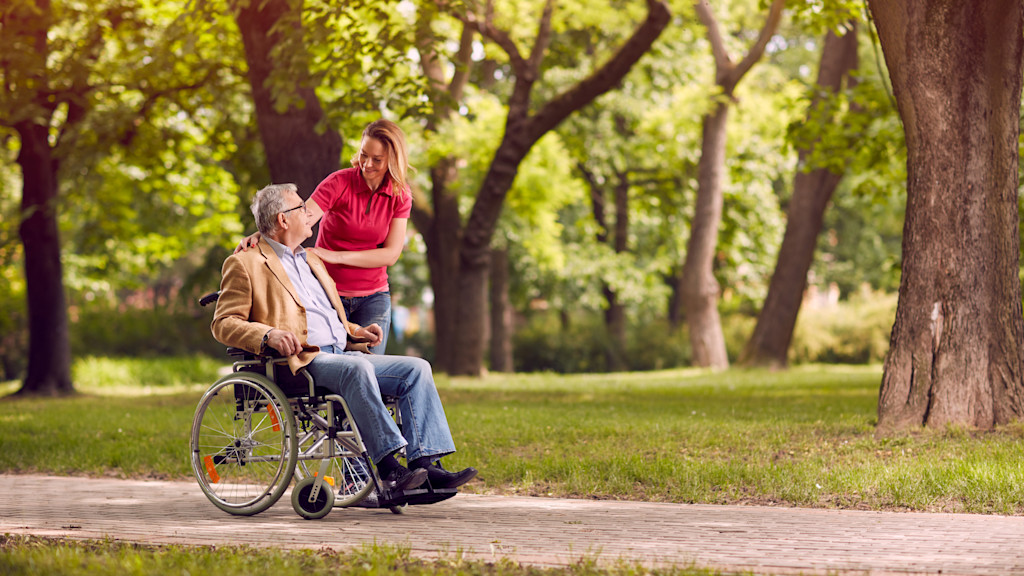 Frau schiebt Rollstuhlfahrer durch einen Park.