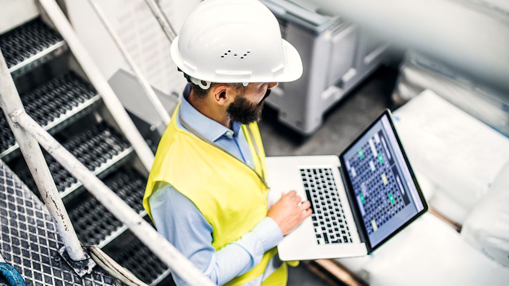 Arbeiter mit Sicherheitshelm und Warnweste arbeitet an Laptop.