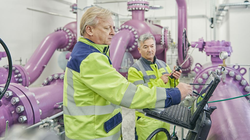 Zwei Techniker stehen zwischen lila Rohren und überprüfen Anlagentechnik.