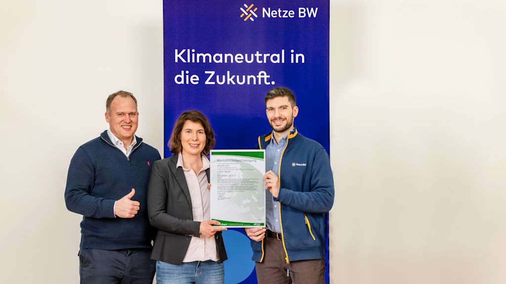 Drei Mitarbeitende der Netze BW präsentieren gemeinsam eine Urkunde zur ISO-Norm 14068-1 Klimaneutralität vor einem Banner mit der Aufschrift ‚Klimaneutral in die Zukunft‘.