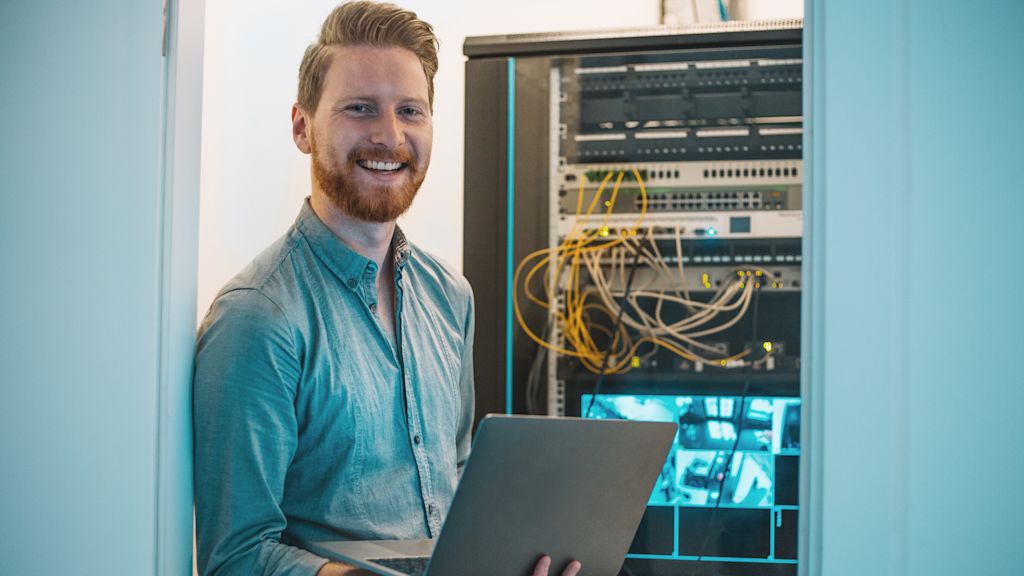 Lächelnder Mann mit Laptop vor Serverrack.