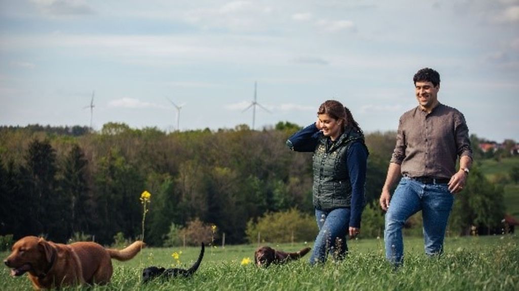 Mann und Frau mit Hunden auf Wiese, Windräder im Hintergrund.