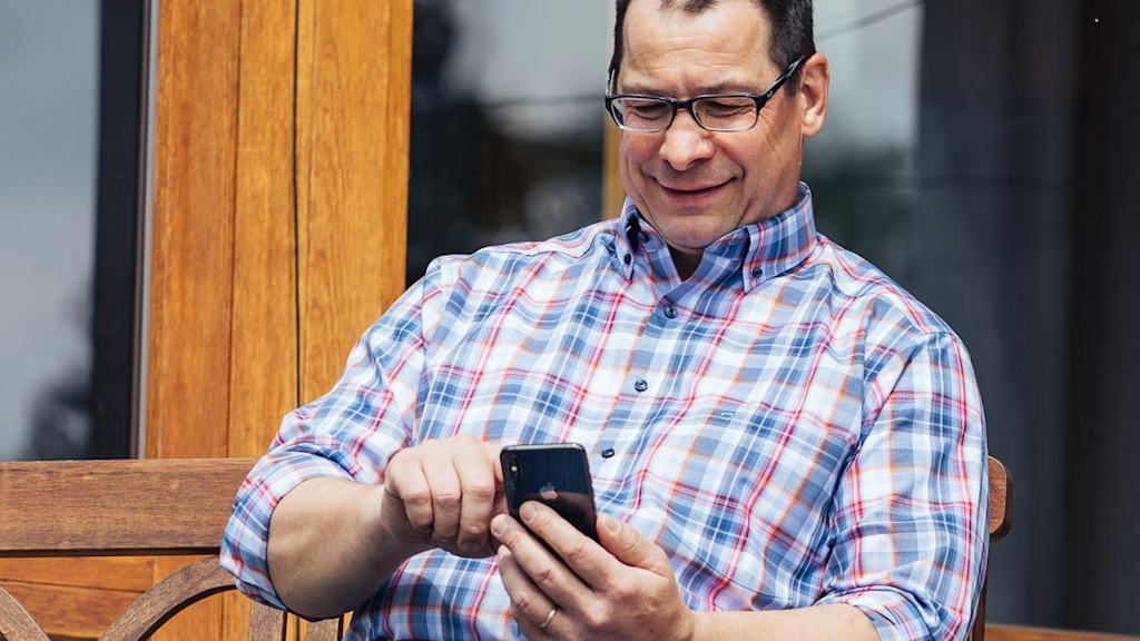 Lächelnder Mann mit Brille benutzt Smartphone vorm Haus sitzend.