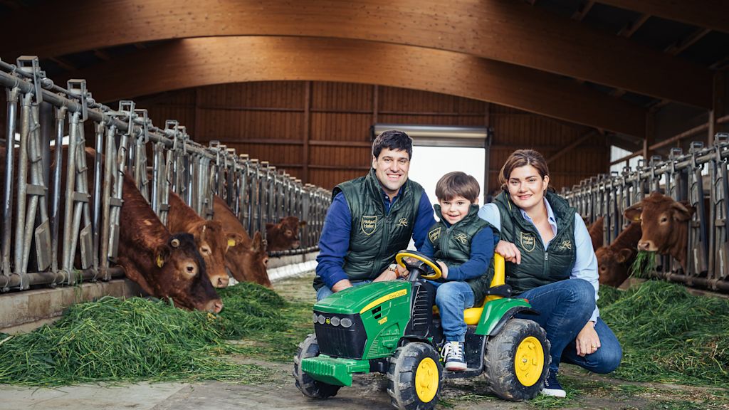 Eine Familie sitzt im Kuhstall vor Rindern am Futtertrog. Ein Kind fährt auf einem kleinen Spielzeugtraktor, die Eltern knien daneben und lächeln in die Kamera.