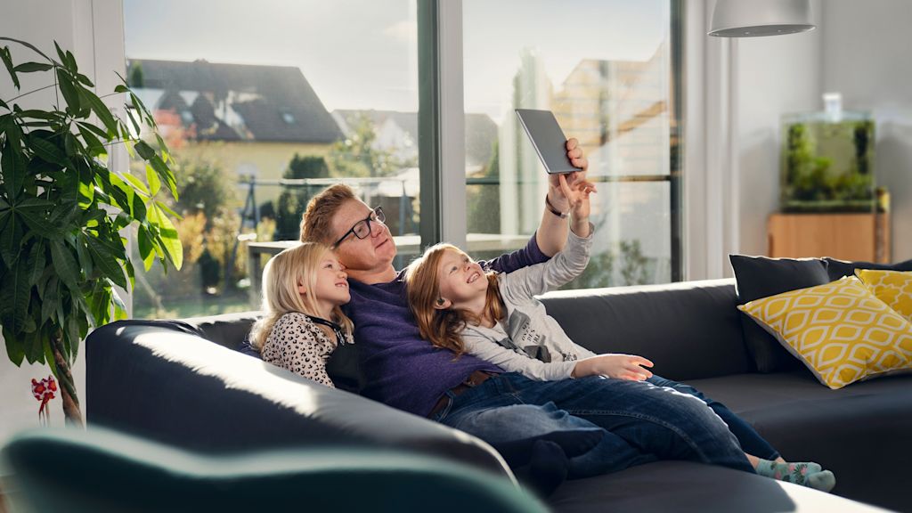 Mann und zwei kleine Mädchen schauen auf dem Sofa gemeinsam auf ein Tablet.