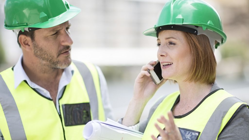 Frau telefoniert auf einer Baustelle, Mann steht daneben und schaut sie an. Beide tragen Sicherheitskleidung.