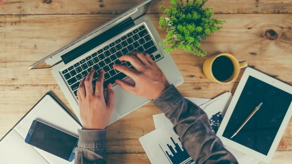 Schreibtisch von oben mit Laptop, Kaffeetasse und Zetteln an dem jemand arbeitet.