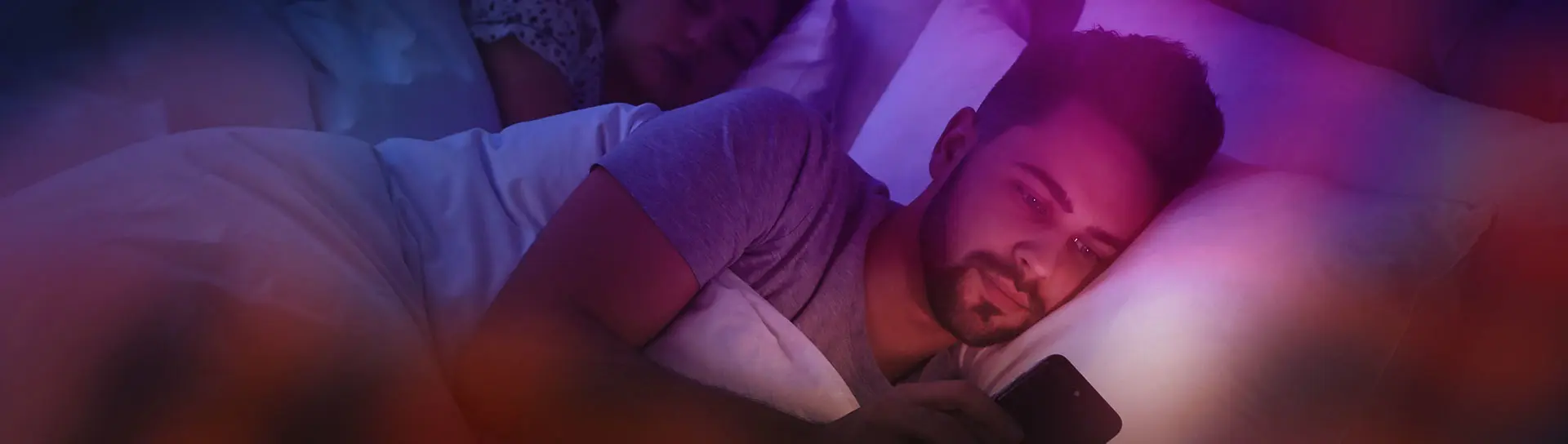 Young man using smartphone while his girlfriend sleeping in bed at night.