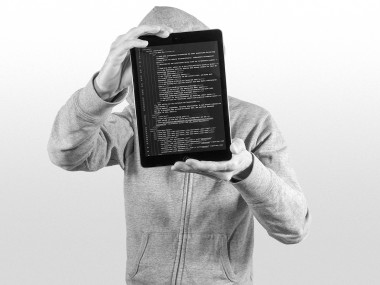 Person mit Kapuzenpullover hält ein Tablet mit Programmcode darauf vor dem Gesicht