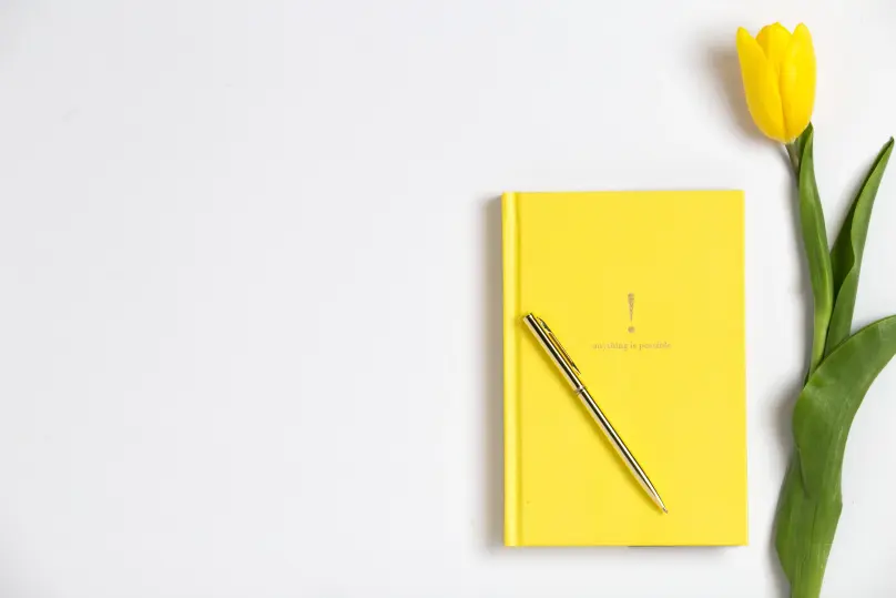 There’s a flat lay of a yellow journal with a gold pen sitting on it. There’s a yellow tulip stem to the right of it. 