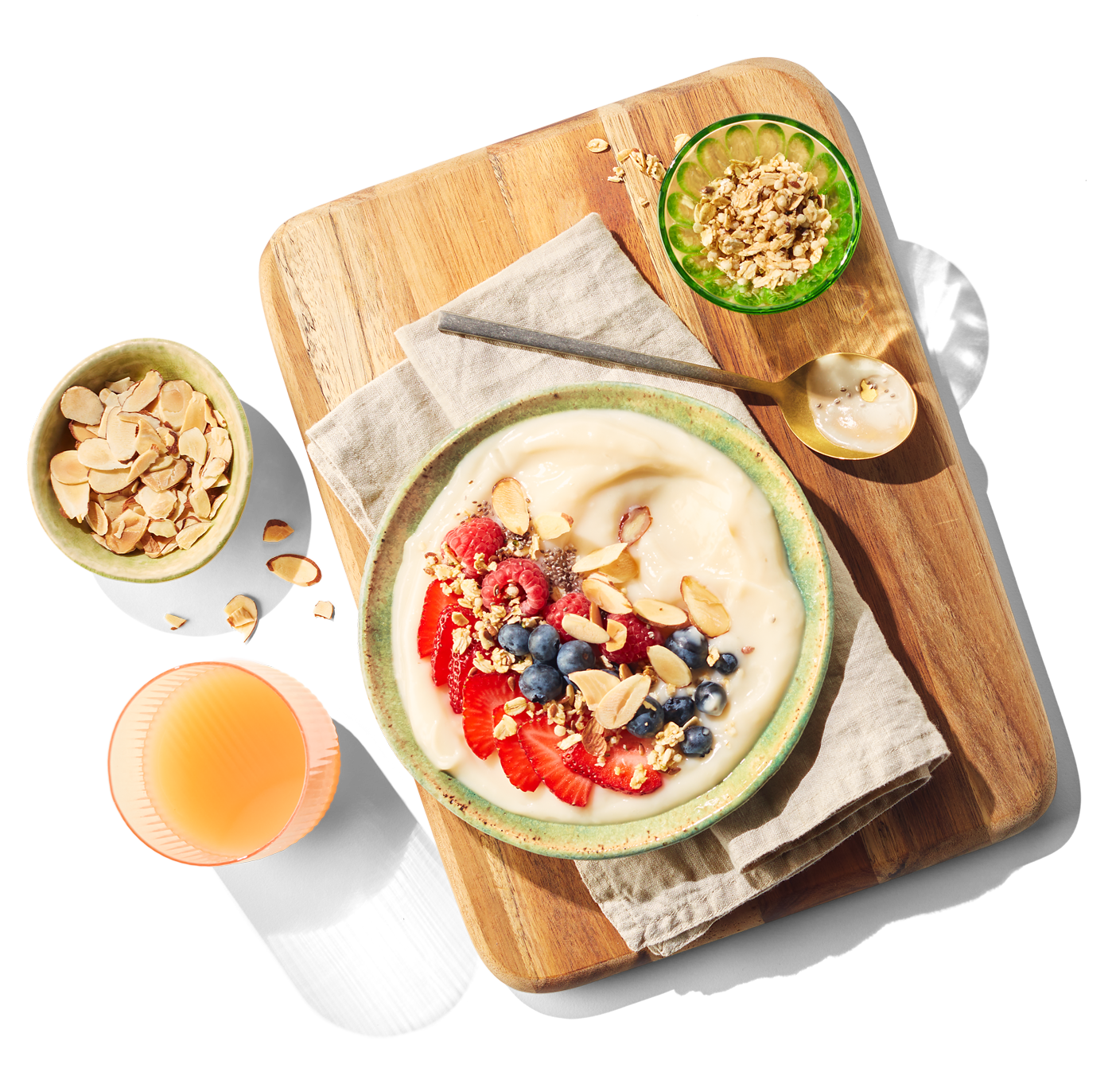 A bowl of plant based yogurt with fruits and oats sitting on a wooden cutting board surrounded by smaller bowls with grains and a glass of orange juice