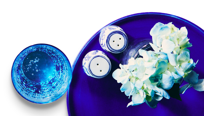 A decorative blue plate with salt and pepper shaker and a pot of flowers on it, beside a glass of water.