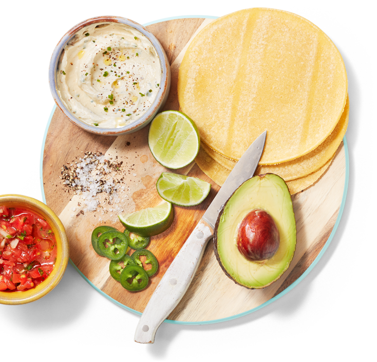 Un bol de trempette au tofu, des tortillas souples, un demi-avocat, des quartiers de lime, des tranches de jalapeno, des assaisonnements et un couteau d’office sur une planche à découper à côté de tomates hachées.