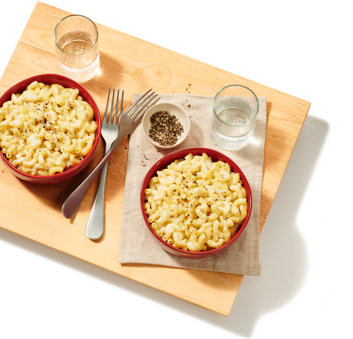 A cutting board with 2 large portions of home made white cheddar mac and cheese, 2 servings of sparkling water and some fresh cracked black pepper on the side