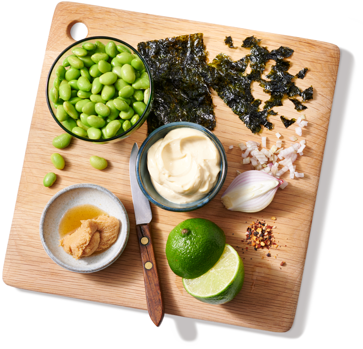 Une planche à découper avec des bols de tartinades et d'edamames, ainsi qu'un peu de nori et d'oignon hachés, une lime coupée en deux et un couteau d'office.