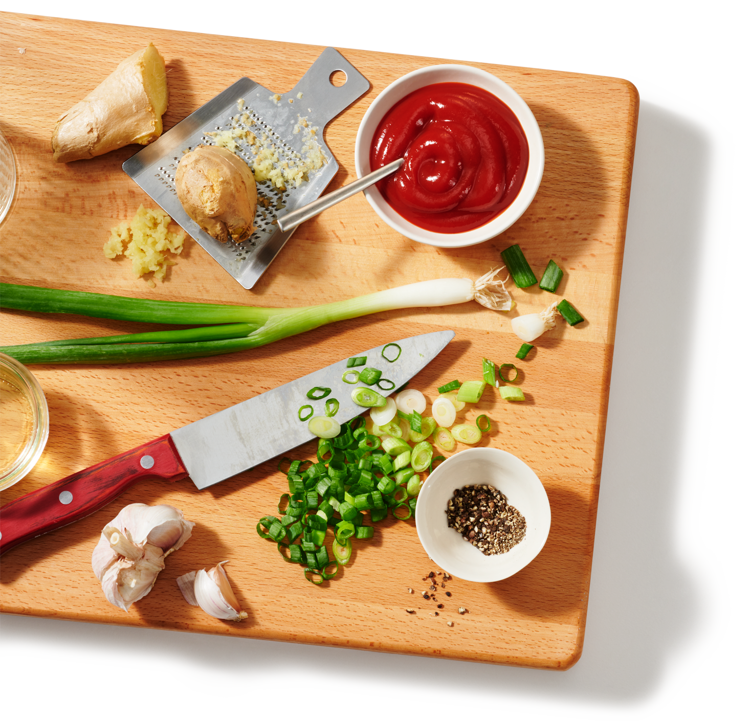 A cutting board with several prepped ingredients to make a spicy stir-fry ketchup chicken dish, including green onions, herbs and a bowl of ketchup