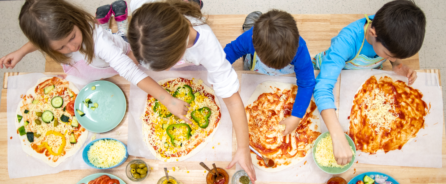 Quatre enfants qui préparent des pizzas à une table en bois dans un cours de cuisine avec des bols de garnitures et de sauces et des assiettes de légumes tranchés.