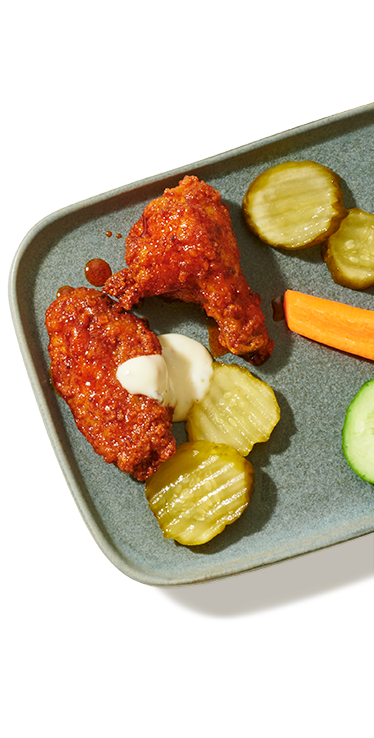 Une portion de deux Ailes de poulet style Nashville PC arrosées d'une sauce crémeuse avec des tranches de pomme de terre, des rondelles de concombre et un morceau de carotte.