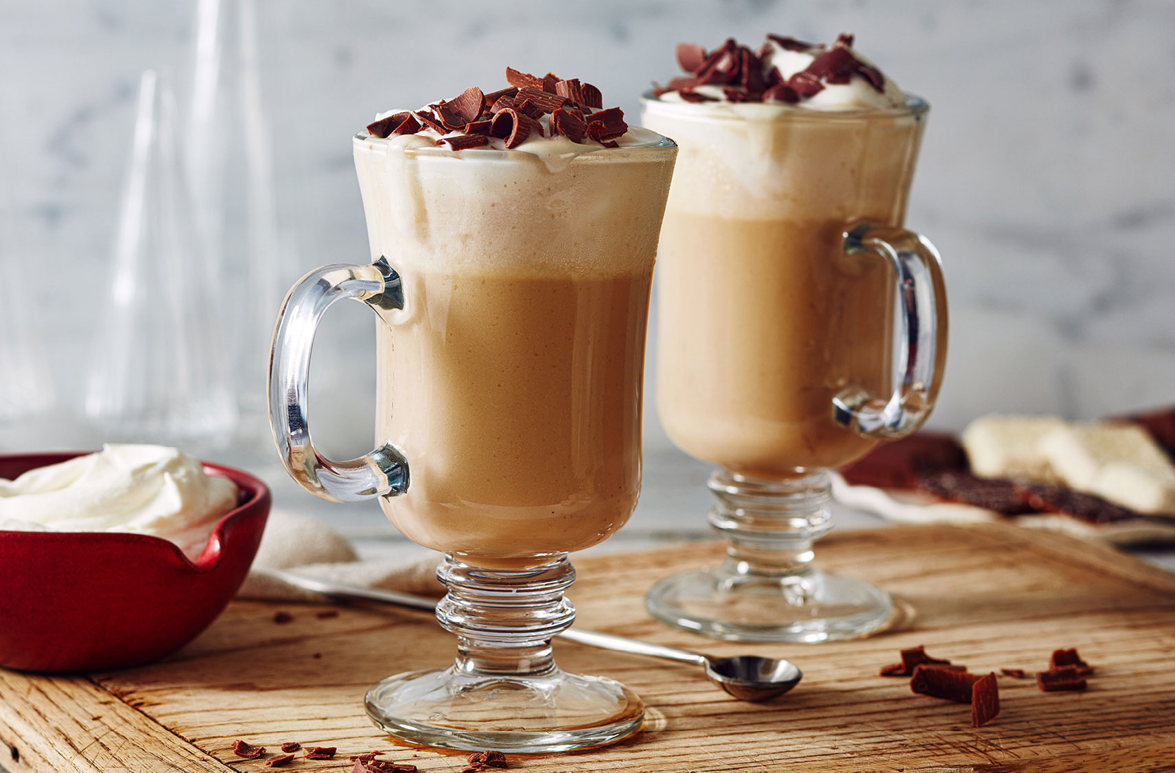 Two warm lattes topped with whipped creamed and chocolate shavings in glass mugs.	