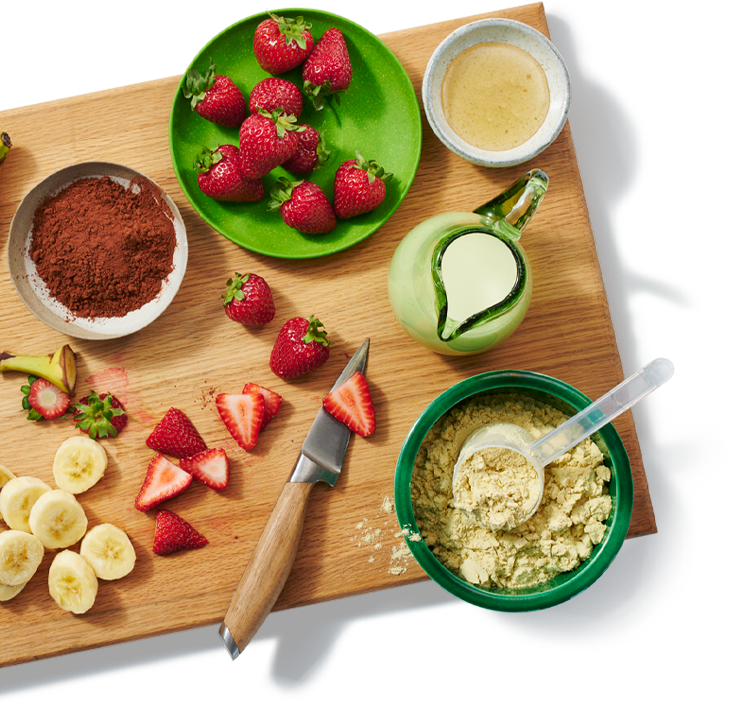 Une planche à découper avec plusieurs ingrédients préparés pour faire un smoothie, comprenant des fraises, des bananes et de la poudre de protéine.