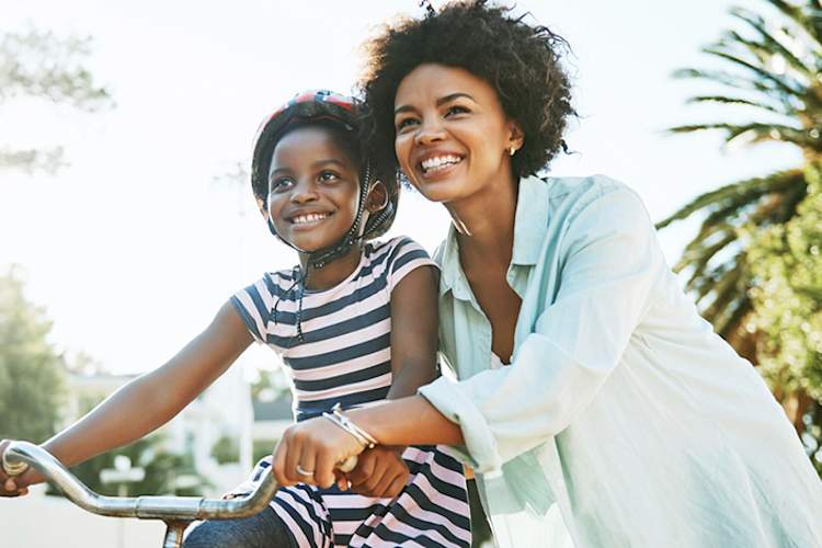 Smiling mother helping her happy young daughter ride a bike outside in the summer.