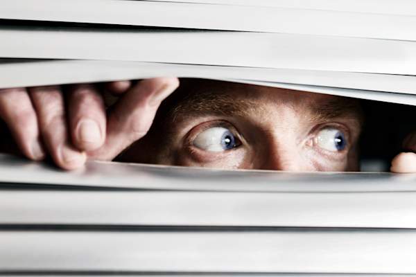 Fearful man looking cautiously through window blinds.