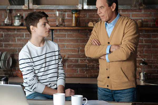 Father talking to teen son in kitchen with open laptop.